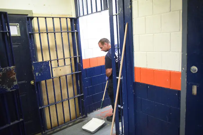 A Corrections staffer is rolling paint onto his paint roller in a cinderblock room that is painted white orange and blue with a jail door
